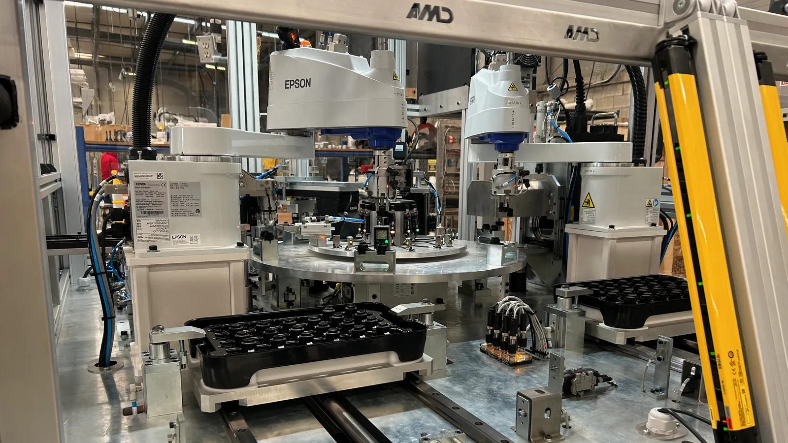 Two Epson SCARA robots with pneumatic grippers over a rotary index table and shuttle-fed parts trays, built by AMD Machines